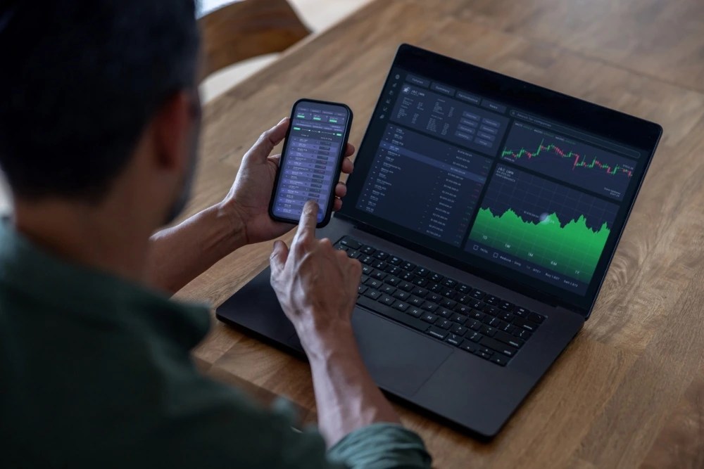 Persona en computador y celular viendo gráficos de inversión
