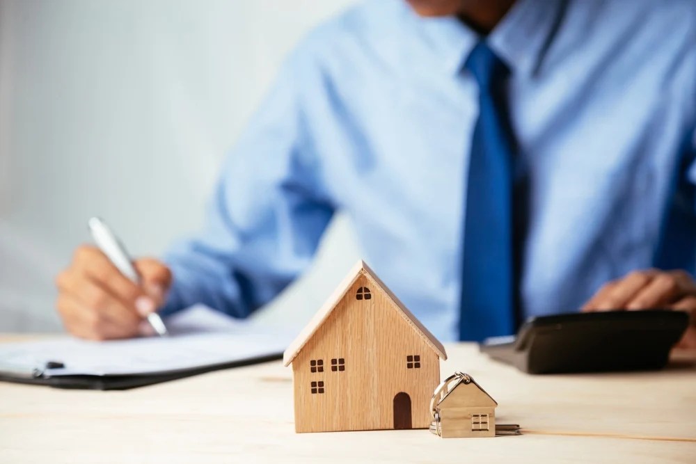 persona escribiendo en un documento de hipoteca, junto a casa miniatura de madera